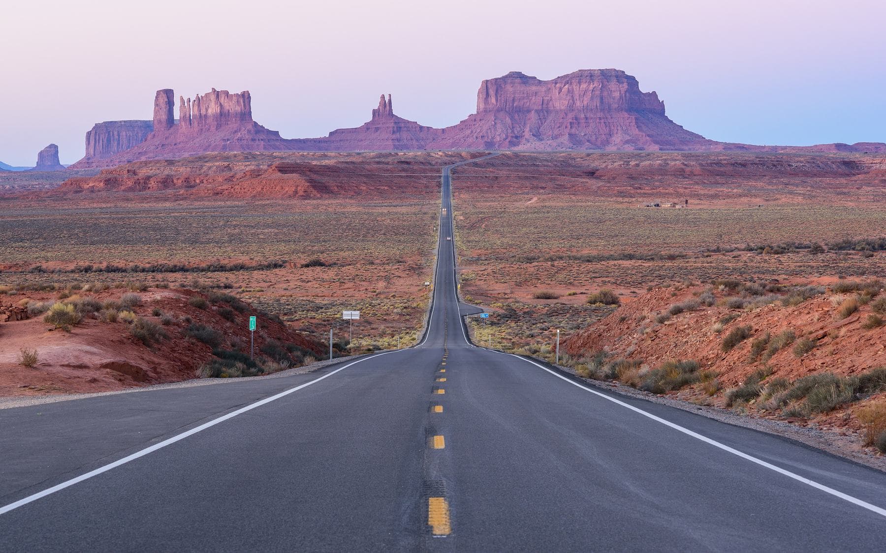 A straight two-lane highway running through red desert toward Monument Valley's sandstone buttes under a pale pink evening sky