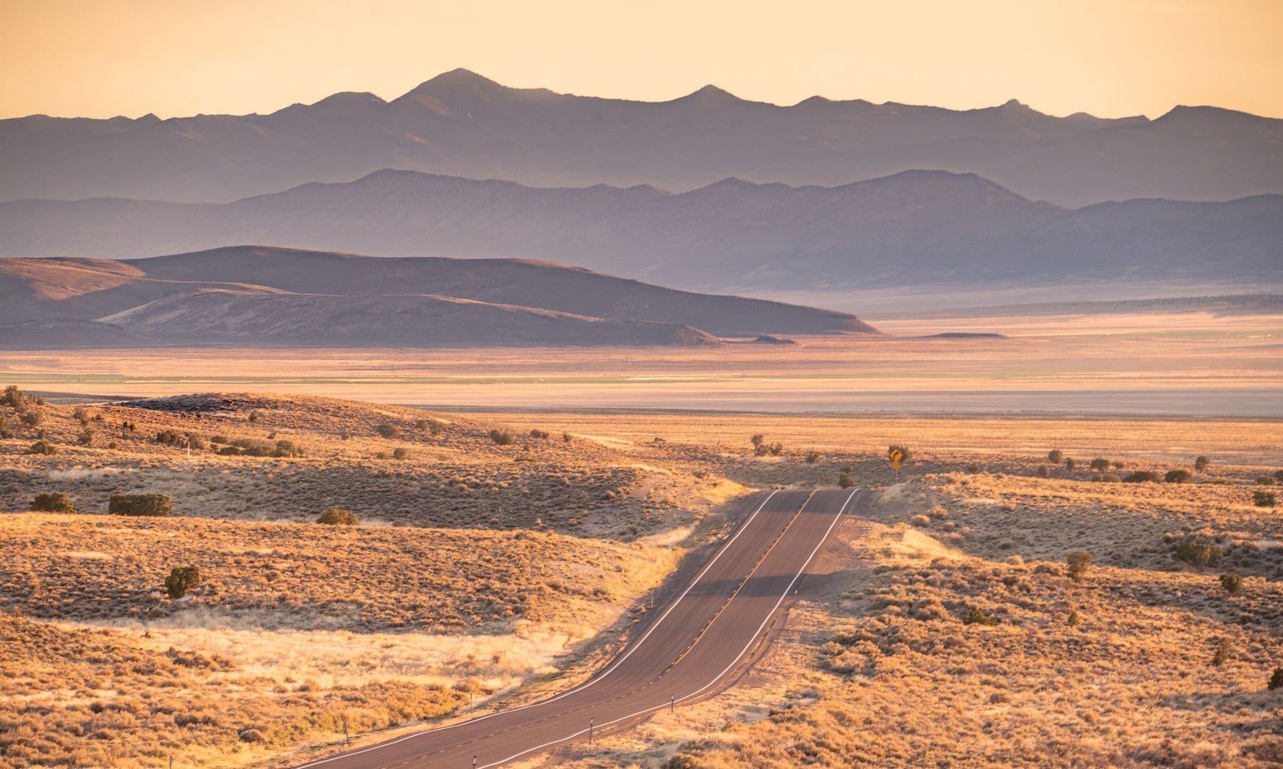A two-lane highway winding through golden sagebrush toward layered mountain ranges in warm evening light